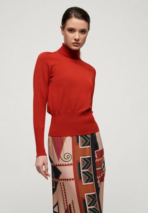 Red turtleneck sweater with long sleeves and ribbed hem, paired with a patterned skirt featuring geometric designs in warm colors.