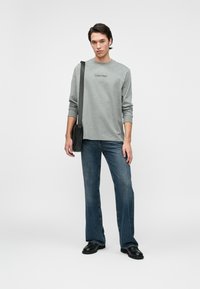 Γκρι μακρυμάνικο μπλουζάκι με λογότυπο Calvin Klein, συνδυασμένο με μπλε τζιν παντελόνι σε καμπάνα. Το μοντέλο κρατάει μια μαύρη τσάντα ώμου με λεία υφή.