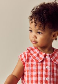 Bambino piccolo con capelli ricci che indossa un vestito a quadri rosso e bianco, guarda di lato su uno sfondo semplice.