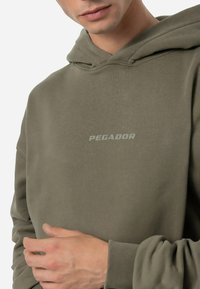 Oliveno zeleni a hoodie iz mehke tkanine, z sprednjo žepom, rebrastimi manšetami in diskretnim logotipom "PEGADOR" v svetlo sivi barvi.
