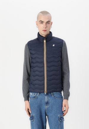 Navy blue quilted vest with high collar, zip closure, and yellow accent. Worn over a gray long-sleeve shirt and paired with denim jeans.