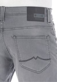 Szare dżinsy z tylną kieszenią z haftowanym wzorem w kształcie chevronu oraz prostokątną czarną metką z napisem "MUSTANG True Denim."