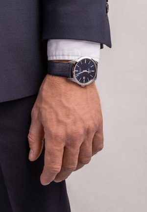 Montre en acier inoxydable argenté avec un cadran bleu marine, des index horaires blancs et un affichage de la date. Associée à un bracelet en cuir bleu foncé.