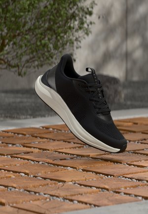 Czarne sportowe buty z białą podeszwą, zawieszone nad drewnianą płytką i kamienistym podłożem, z rozmytym drzewem i ścianą w tle.