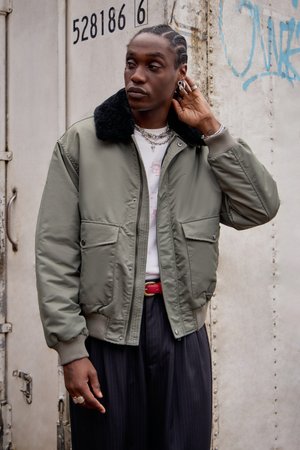 Blouson bomber vert olive avec un col en fourrure noire, deux poches avant, des poignets côtelés, porté par-dessus une chemise blanche et un pantalon à rayures.