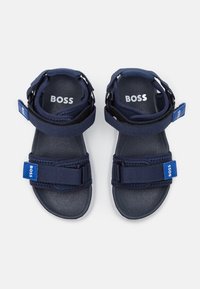 Tummansiniset sandaalit kangasremmeillä ja tarranauhafastauksilla. Varustettu teksturoidulla pohjalla ja valkoisella kumipohjalla. "BOSS"-logo remmeissä.