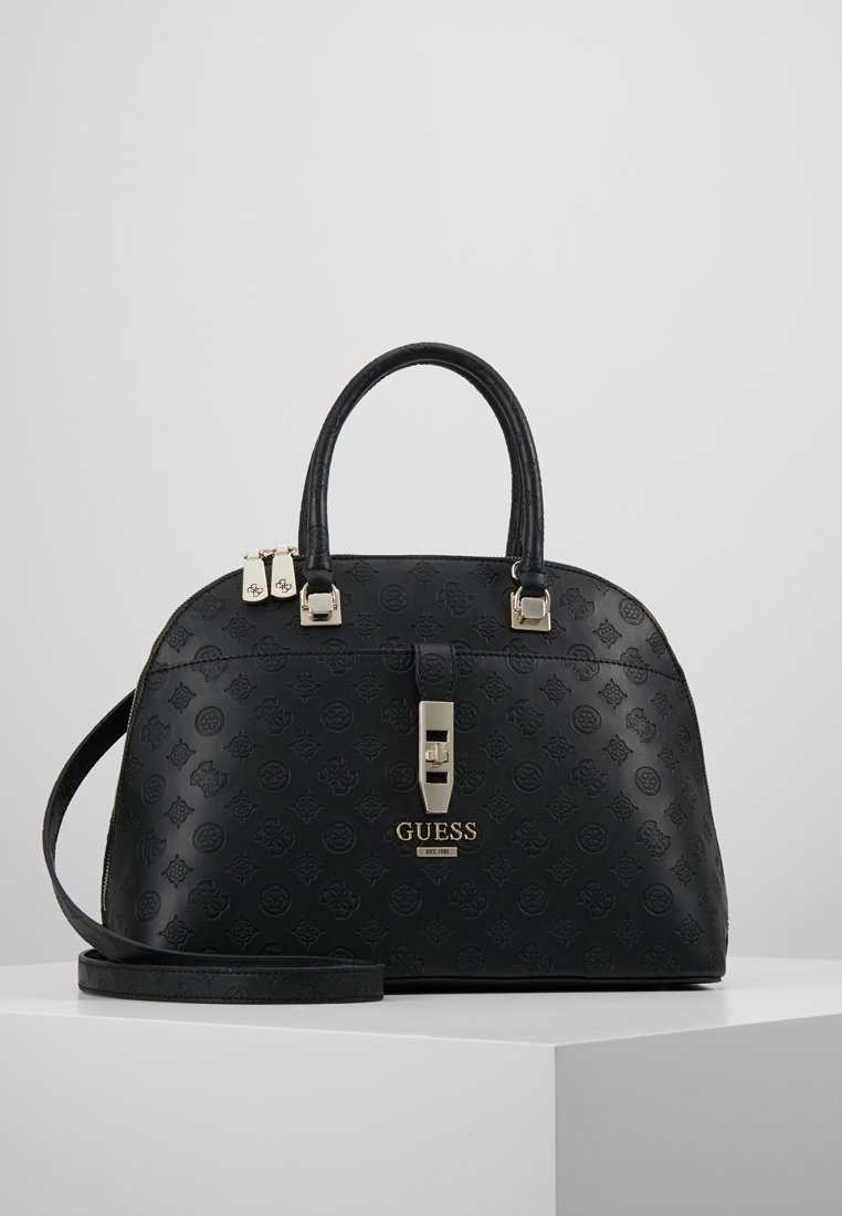Sac à main noir Guess avec motif embossé, doubles poignées, bandoulière réglable, fermeture à serrure frontale et plaque logo en métal sur une surface blanche.