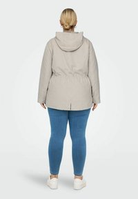 Bežna pufasta jakna s kapuco, elastičnim pasom in zadnjim delom za zračenje. Model nosi modre oprijete kavbojke in bele športne copate.