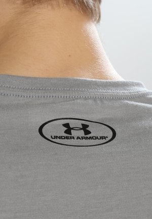 Nærbillede af bagsiden af nakkeområdet på en person iført en lysegrå skjorte med en sort Under Armour-logo og tekst under kraven.