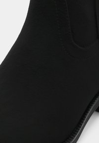 Botim preto até ao tornozelo com acabamento em camurça texturizada, ponta arredondada e detalhes de costura nas laterais. Apresenta um tacão baixo e modesto.