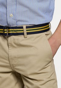 Personne portant un pantalon beige avec une ceinture rayée bleu marine, jaune et verte, associé à une chemise rayée bleu et blanc rentré, main dans la poche.