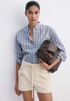 Donna che indossa una camicia a quadri blu e pantaloncini beige tiene una grande pochette in pelle marrone su sfondo bianco.