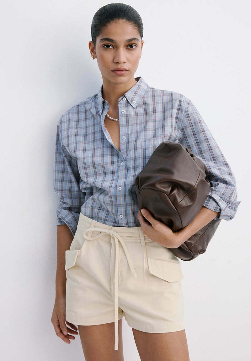 Femme portant une chemise à carreaux bleue et un short beige tient une grande pochette en cuir marron sur un fond blanc.