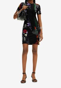 Figurbetontes schwarzes Kleid mit mehrfarbigen geometrischen Mustern, rundem Ausschnitt und kurzen Ärmeln, kombiniert mit einer schwarzen Handtasche und Riemchensandalen.