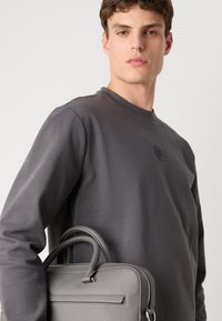 Gray leather handbag with twin handles, textured finish, and a zip closure. Worn with a matte gray long-sleeve shirt featuring a logo.