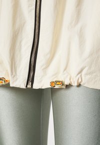 Chaqueta cremallera de un tono beige claro con cremallera negra, dobladillo ajustado con herrajes dorados, combinada con leggings metálicos verdes. Textura suave, diseño casual.