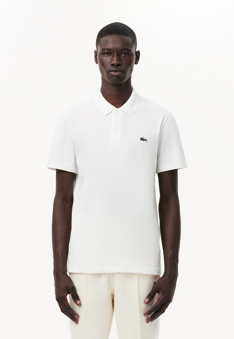 Biely polo tričko s klasickým golierom a krátkymi rukávmi, s malým námorníckym logom Lacoste na ľavej hrudi. Hladká, ľahká tkanina.