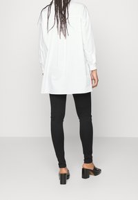 Top blanco de manga larga con un dobladillo curvado, combinado con leggings negros ajustados y mules de tacón bajo negros. La tela parece suave y casual.
