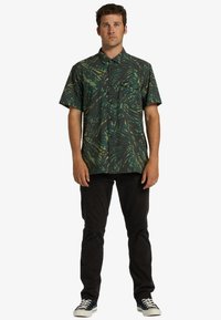 Short-sleeve shirt with a dark background and green abstract leaf pattern, featuring a button-up front and a chest pocket. Worn with black pants.