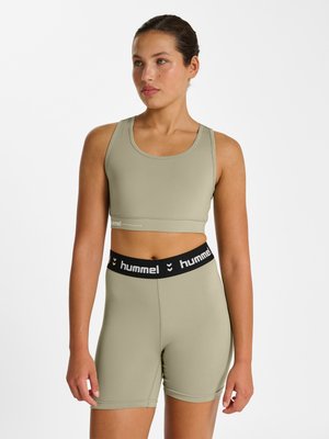 Jonge vrouw draagt een lichtgroene sportbeha en bijpassende shorts met hoge taille met zwarte "hummel" tailleband, staand tegen een witte achtergrond.