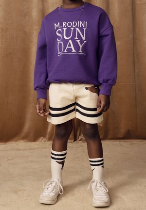 Sweat-shirt violet avec l'inscription « M. Rodini SUN DAY » en lettres argentées, short crème avec des rayures noires, chaussettes blanches à rayures et baskets blanches.