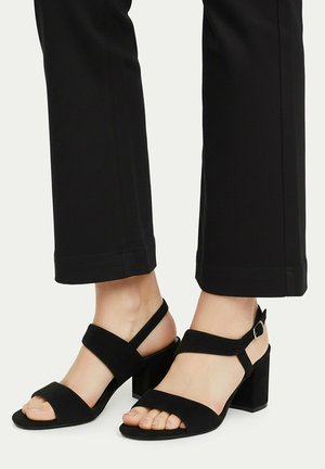 Füße in schwarzen Sandalen mit offenem Zehenbereich und Blockabsätzen, die mit Knöchelriemen versehen sind, kombiniert mit schwarzen, gekürzten Hosen vor einem hellen Hintergrund.