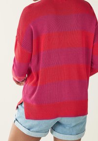 Pull-over en rayures horizontales rouge vif et rose, avec texture côtelée, fentes latérales et épaules tombantes.