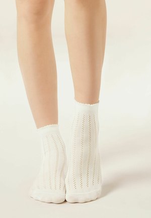 Jambes portant des chaussettes blanches montantes avec des motifs à rayures verticales en maille ajourée sur un fond neutre.