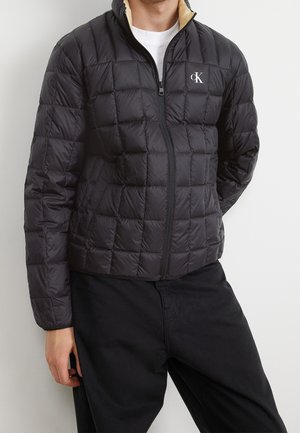 Schwarze Daunenjacke mit gestepptem Design und hohem Kragen, ausgestattet mit einem beigen Innenfutter, einem Reißverschluss vorne und einem kleinen Logo auf der Brust.