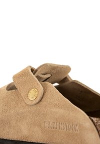 Kadar blizu bež antilop Birkenstock klompe s podesivim remenom i metalnim gumbom na bijeloj pozadini.