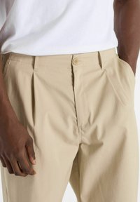 Beige cotton trousers with pleats, button closure, and side pockets. Smooth texture, straight-leg design, styled with a white shirt.