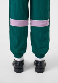 Calças de jogging verdes e leves com bandas contrastantes em lavanda e punhos elásticos, combinadas com sapatos pretos e meias brancas. Tecido texturizado plano.