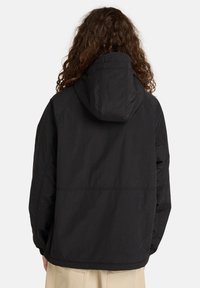 Person mit langen lockigen Haaren, die einen schwarzen Kapuzenjacke trägt und von hinten zu sehen ist, kombiniert mit beigen Hosen vor einem schlichten weißen Hintergrund.