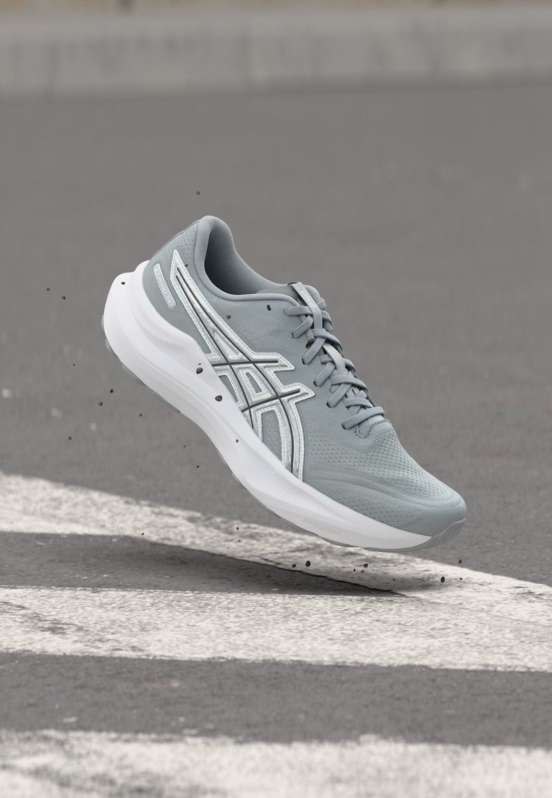 Ανοιχτό γκρι αθλητικό παπούτσι με λευκή σόλα και πλευρικές ρίγες, στον αέρα πάνω από διάβαση πεζών σε άσφαλτο με μικρά υπολείμματα γύρω.