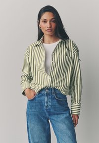 Camicia a righe verdi e bianche con bottoni e taschino, indossata sopra una maglietta bianca e abbinata a jeans blu a vita alta.