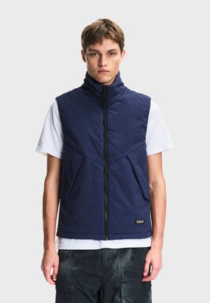 Jeune homme portant un gilet matelassé sans manches bleu marine par-dessus un t-shirt blanc et un pantalon cargo foncé, debout devant un fond clair uni.