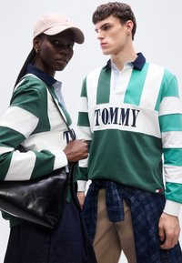 Δύο μοντέλα φορούν πράσινα και λευκά ριγέ πουκάμισα Tommy· το ένα κρατά μια μαύρη δερμάτινη τσάντα και φοράει μπεζ καπέλο.