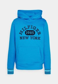 Felpa blu con tasca frontale, cappuccio con cordino, testo ricamato "HILFIGER NEW YORK 1985" e dettagli a strisce sui polsini.