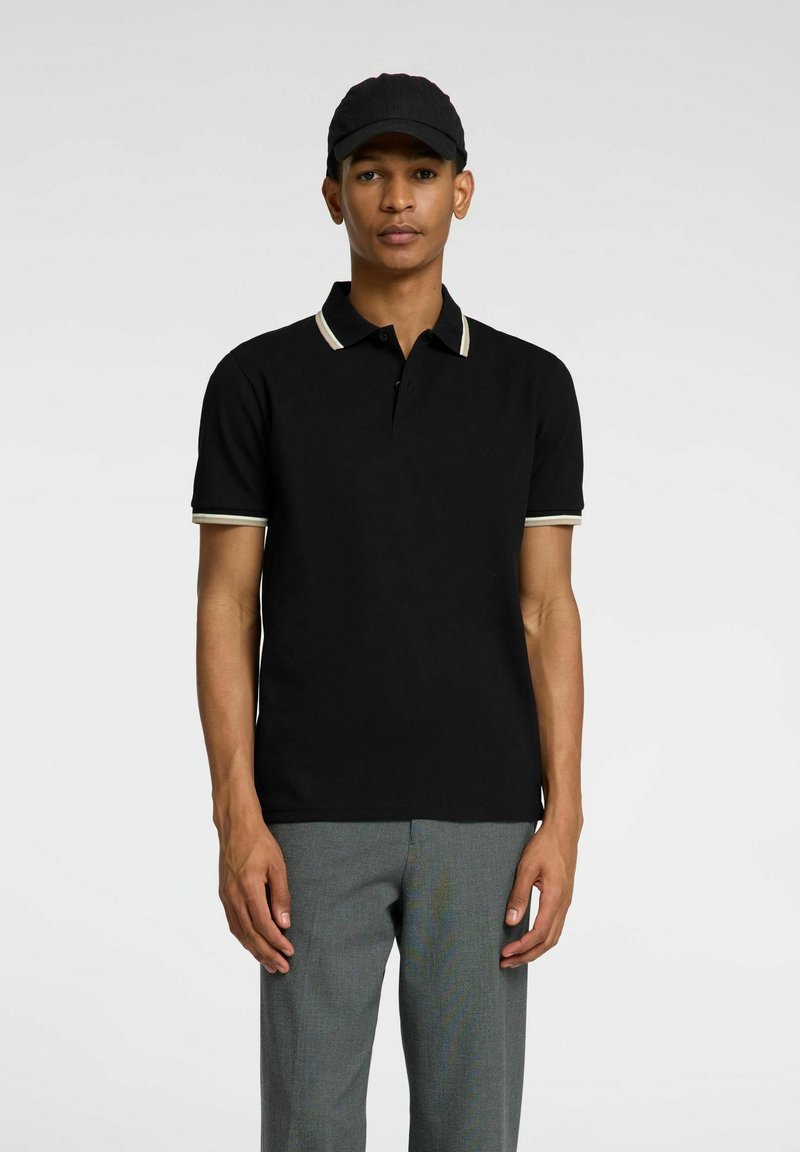 Polo shirt noir à manches courtes avec col et bord des manches beige contrastants, en tissu mélangé de coton, associé à un pantalon gris.