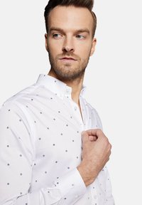 Biała koszula z zapięciem na guzik w kołnierzyku, ozdobiona małymi czarnymi gwiazdkami, dopasowana w kształcie, z długimi rękawami. Gładka tekstura, minimalistyczny design.