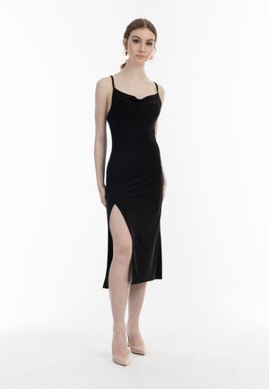 Junge Frau in einem schwarzen ärmellosen Kleid mit hohem Beinschlitz, trägt nudefarbene High Heels und auffällige Ohrringe, steht vor weißem Hintergrund.