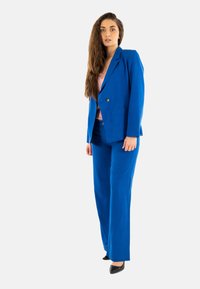 Chaqueta y pantalones de traje azul con un corte relajado, con cierre de un solo botón y textura de tela suave. Llevado sobre una blusa de color rosa claro.