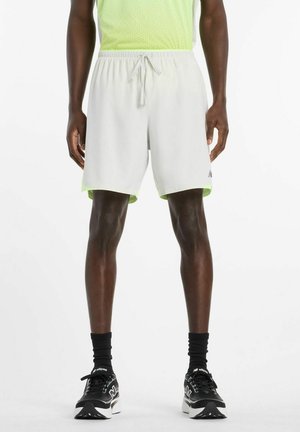 Man draagt lichtgrijze sportshorts met trekkoord, zwarte sokken en zwarte hardloopschoenen, staand tegen een witte achtergrond.