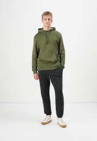 Young man standing wearing an olive green hoodie, dark gray joggers, and white sneakers against a plain white background.
