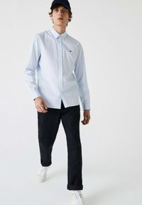 Lichtblauw button-up overhemd van een katoenmix stof, met een kraag, lange mouwen en een klein logo op de borst, gecombineerd met donkere broeken en witte sneakers.