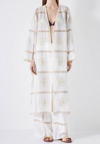 Weißer bestickter Kaftan mit floralen Mustern und transparenter Textur, mit langer Silhouette und offener Vorderseite. Kombiniert mit lockeren weißen Hosen.