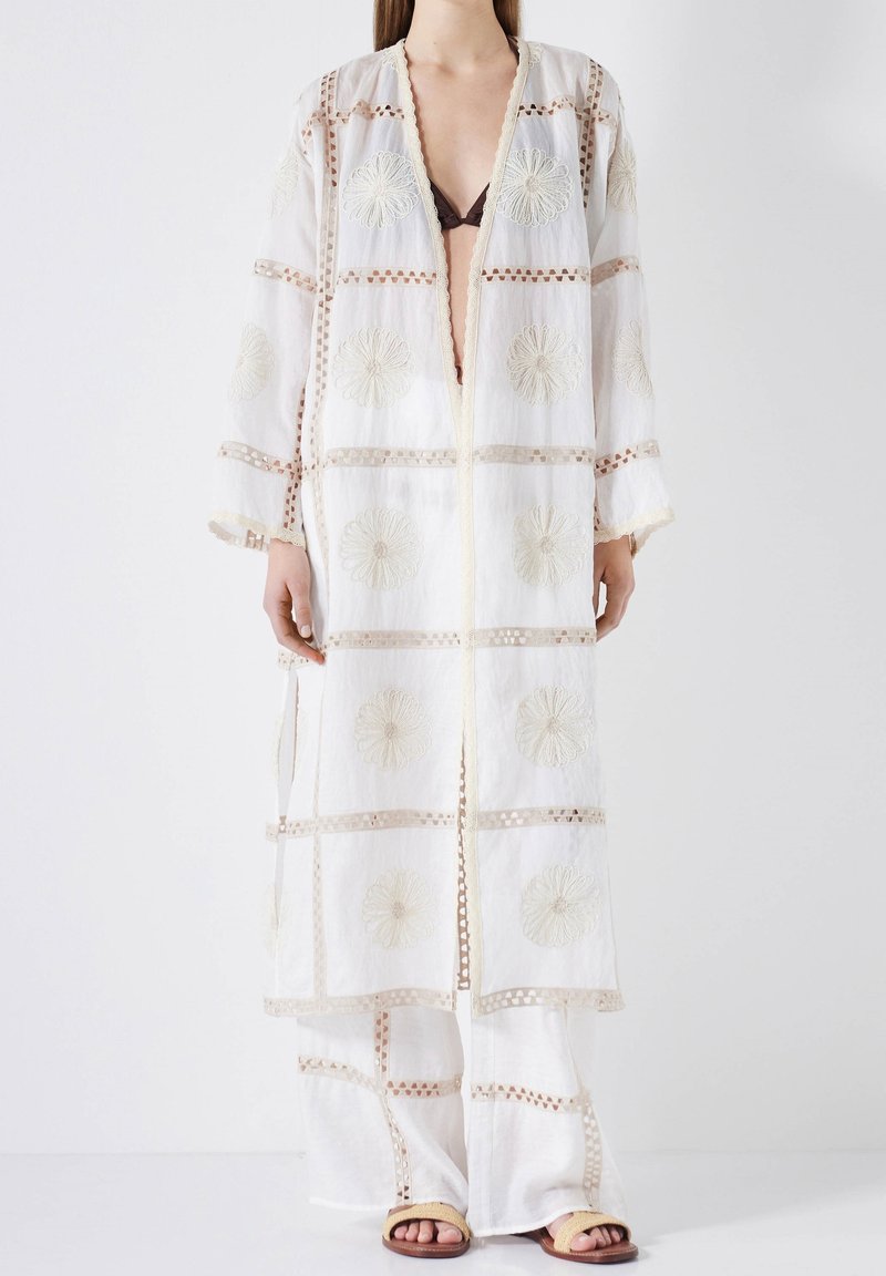 Weißer bestickter Kaftan mit floralen Mustern und transparenter Textur, mit langer Silhouette und offener Vorderseite. Kombiniert mit lockeren weißen Hosen.