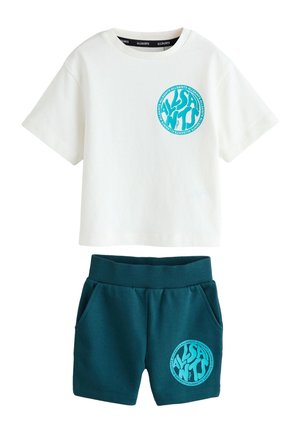 Ung dreng med kort hår iført en hvid t-shirt med blå AllSaints-logo på ryggen og mørkegrønne shorts står med hænderne i lommerne.