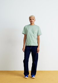 T-shirt in cotone verde chiaro con maniche corte e un piccolo logo dorato sul petto, abbinato a pantaloni in denim blu scuro. Le sneakers sono blu e bianche.