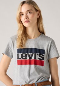 T-shirt en coton gris avec un grand logo Levi's en bleu marine et rouge, sur un fond gris uni. Col rond et manches courtes.